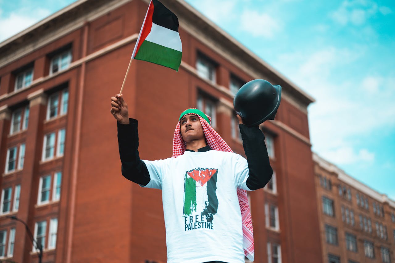 Man waving a Palestine flag in protest with an urban backdrop.